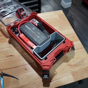 May include: A red plastic storage case with a clear lid containing a grey NOCO Boost HD jump starter, red and black jumper cables, and a grey internal divider. The lid features a circular sticker with the text 10 SPOT TOOLS.