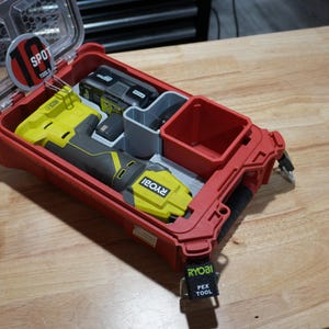 May include: A red plastic tool storage case with a clear hinged lid, containing a yellow and gray Ryobi power tool and a battery. The case features internal compartments, a circular 10 SPOT TOOLS label, and a black tag labeled RYOBI PEX TOOL.