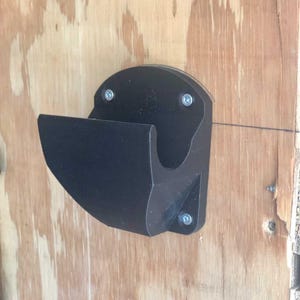 Puede incluir: Un soporte negro montado en la pared, posiblemente para una manguera u otro artículo, está fijado a una superficie de madera con cuatro tornillos. El soporte tiene un diseño curvo con una parte superior semicircular y un frente curvo.