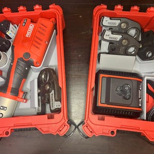 以下が含まれることがあります： 2つの開いた赤いツールケース。配管工具が入っています。1つのケースには、Ridgid RP 241プレスツール、継手、パイプが入っています。もう1つのケースには、プレスツールジョー、充電器、バッテリーが入っています。