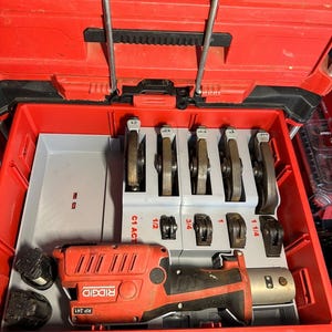 May include: Red tool case with a red and black Ridgid RP 241 press tool and various press fitting jaws. The case is open, revealing the tool and jaws in a gray tray. The jaws are labeled with sizes.