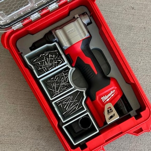 Op de afbeelding: Een rode en zwarte Milwaukee M12 klinknagelgereedschapskit in een rode koffer. De kit bevat het gereedschap, klinknagels en een opbergvak. Het gereedschap heeft een zilveren kop en een zwarte handgreep. Het Milwaukee-logo is zichtbaar.