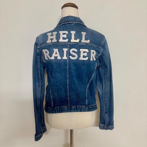 May include: A blue denim jacket with the words "HELL RAISER" in silver glitter lettering across the back. The jacket is displayed on a mannequin against a white background. The jacket has a classic collar and button-down front.