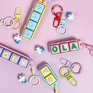 Może przedstawiać: Kolekcja kolorowych prostokątnych breloków z mechanicznymi przełącznikami klawiaturowymi jako przyciskami. Breloki zawierają imiona takie jak DOUG, OLA i JOE oraz symbole, takie jak gwiazdka i kwiatek, przymocowane do różnych metalowych karabińczyków.