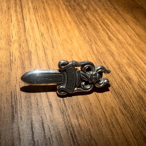 May include: A silver-colored dagger-shaped pendant with ornate scrollwork and the word "Chrome" engraved on the handle. The pendant has a pointed blade and is displayed on a wooden surface.