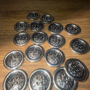 May include: A collection of silver-colored buttons with an ornate floral design. Each button has four holes and a raised, decorative pattern around the edges. The buttons are scattered on a wooden surface.
