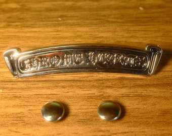 Puede incluir: Una placa de metal plateado con el texto "CHROME HEARTS" en relieve en una fuente gótica estilizada, acompañada de dos botones o tachuelas de metal plateado, pequeños, lisos y circulares, situados debajo de la placa.