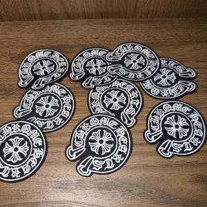 Può includere: Una collezione di nove toppe ricamate circolari in bianco e nero. Ogni toppa presenta un emblema a croce centrale circondato dalla scritta Chrome Hearts in carattere gotico, disposta in un motivo a bordo circolare con una piccola icona a pugnale.