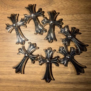 May include: Seven silver-colored cross pendants arranged in a circular pattern. Each cross has a detailed, ornate design with rounded edges and a central stud. The crosses are displayed on a wooden surface.