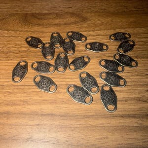 May include: A collection of small, oval-shaped metal charms with a circular design in the center and two holes on either end. The charms are silver in color and are scattered on a wooden surface.