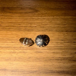May include: Two silver buttons with a decorative design. One button has a loop on the back, the other has a domed, floral-like pattern. The buttons are set against a wooden background.