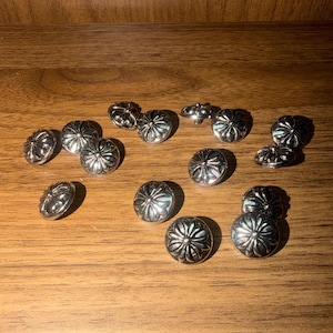 Può includere: Una collezione di tredici bottoni metallici circolari color argento, ognuno caratterizzato da un motivo floreale o a stella in rilievo con petali radianti che si estendono da un punto centrale, disposti in modo sparso su una superficie piana.