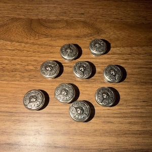 May include: A collection of eight circular silver-toned metal buttons, each featuring a raised central cross emblem surrounded by intricate decorative scrollwork and engraved detailing on the outer rim, arranged in a scattered formation.