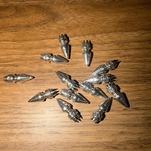 May include: A collection of small, silver-colored metal charms shaped like stylized carrots with crown-like tops. The charms are scattered on a wooden surface, with varying orientations and angles.