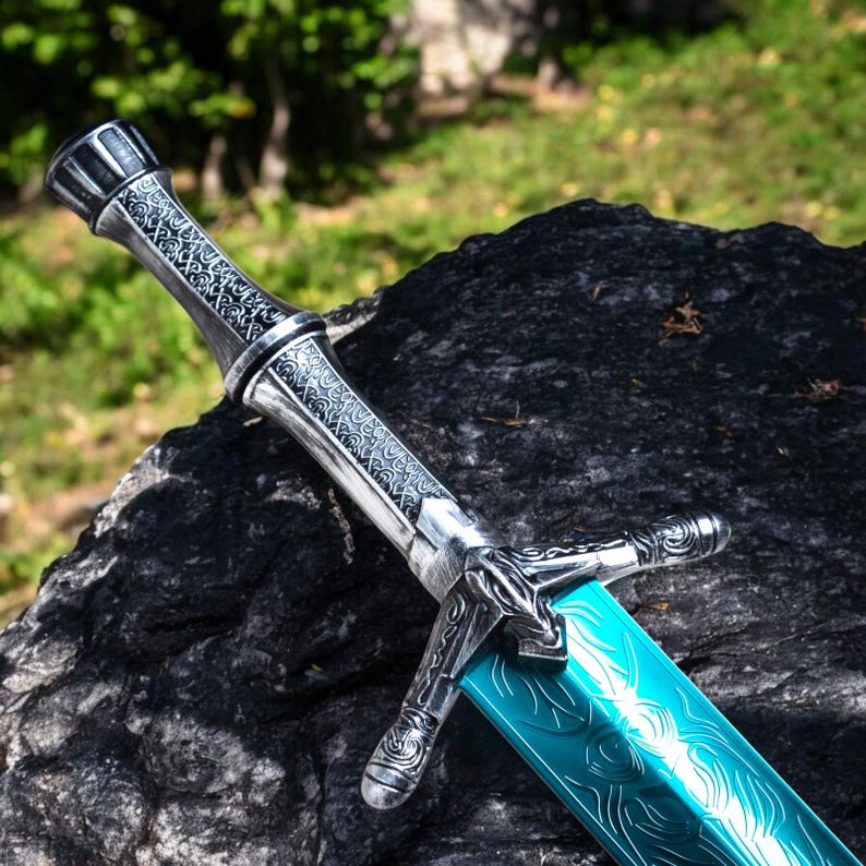May include: A detailed sword with a silver handle and a turquoise blade. The handle features intricate patterns and a decorative pommel. The blade has a wave-like design etched into its surface. The sword is displayed on a dark, textured rock.