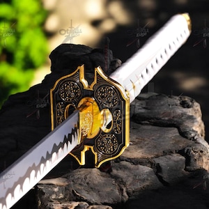 Puede incluir: Primer plano de una katana. El mango es blanco, la hoja plateada y la guarda dorada y negra con un patrón intrincado. La espada se exhibe sobre una superficie oscura y texturizada, resaltando los detalles.