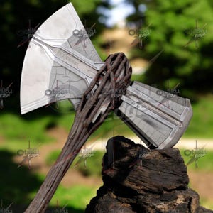 May include: A detailed replica of a double-bladed axe, featuring a silver-colored head with intricate detailing and a wooden handle. The handle is wrapped with a textured, dark brown material. The axe rests on a dark, textured surface.