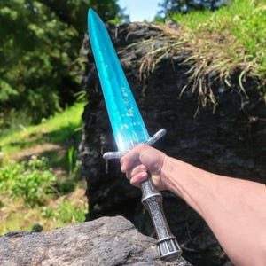 May include: A fantasy sword with a translucent, light blue blade and a silver-colored hilt. The blade has etched text. The hilt features intricate designs and a decorative pommel. The sword is held against a natural outdoor background.