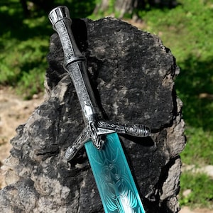 May include: A decorative sword with a silver handle and a blue blade. The handle features intricate designs, and the blade has a patterned surface. The sword is displayed against a dark, textured background, suggesting a fantasy or historical theme.