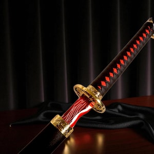 May include: A decorative sword with a black handle wrapped in red diamond-patterned stitching. The blade is red and white, with gold accents on the hilt and pommel. The sword is displayed on a dark surface.