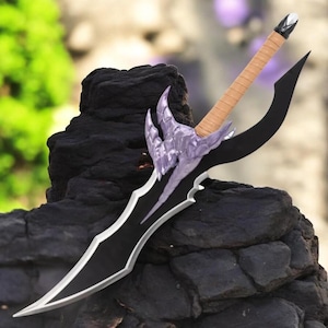 May include: A black and silver fantasy sword with a curved blade and ornate purple hilt. The handle is wrapped in tan material. The sword is displayed on a dark, textured surface, with a blurred green and tan background.