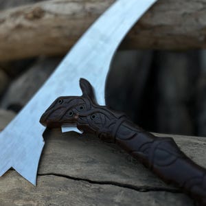 May include: A close-up of a curved blade with a dark brown, intricately carved handle. The blade is silver and has a sharp edge. The handle features detailed knotwork and a stylized animal head.
