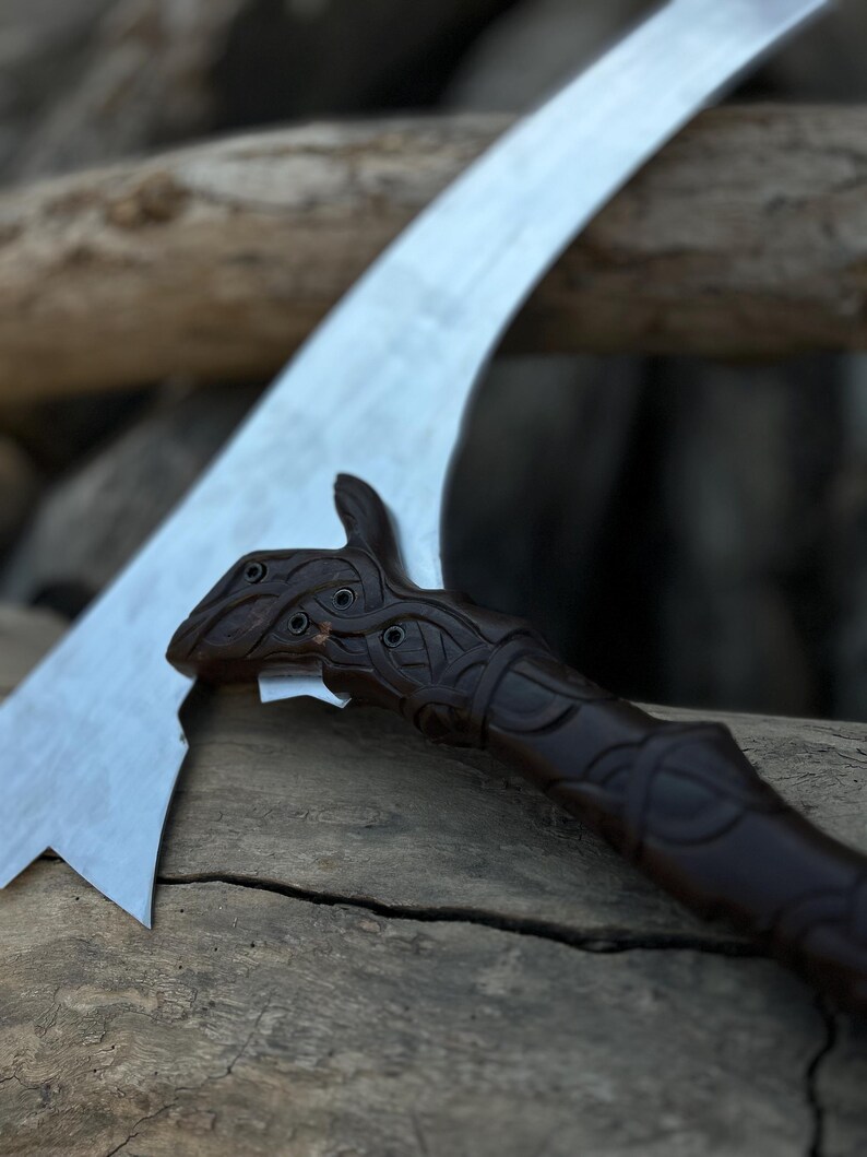 May include: A close-up of a curved blade with a dark brown, intricately carved handle. The blade is silver and has a sharp edge. The handle features detailed knotwork and a stylized animal head.