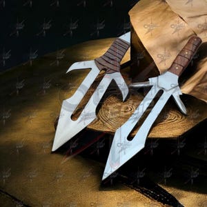 Könnte beinhalten: Zwei silberne und braune Wurfmesser mit einzigartigen Klingendesigns. Die Klingen haben eine markante Form mit einem zentralen Punkt und gebogenen Kanten. Die Griffe sind mit braunem Leder und Holz umwickelt. Die Messer werden auf einer Holzoberfläche präsentiert.