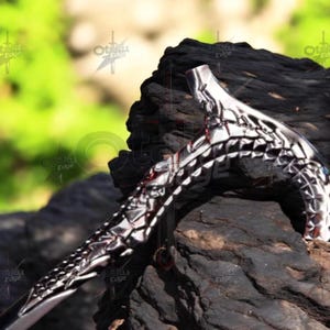 May include: A silver and black decorative sword with a detailed geometric design. The sword is resting on a dark, textured surface, with a blurred green background.