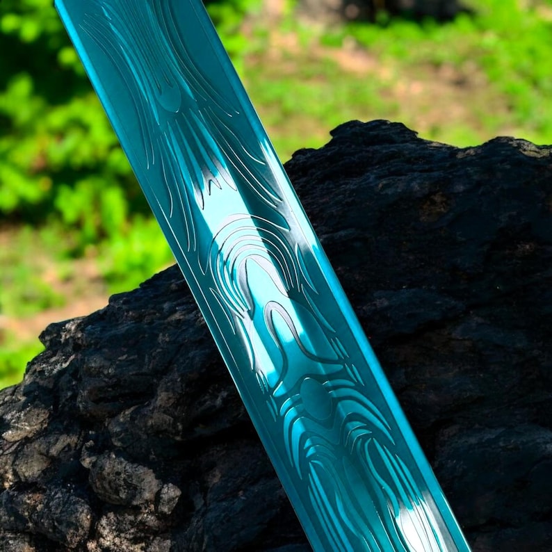 May include: Close-up of a teal-colored sword blade with intricate etched patterns. The blade has a reflective surface and is set against a dark, textured background with a blurred green backdrop. The design suggests a fantasy or collectible item.