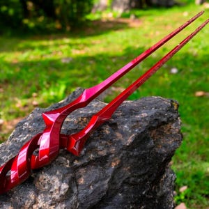May include: A pair of glossy, red-colored scissors with a unique, stylized design. The scissors are resting on a dark gray rock, with a blurred green background of foliage.