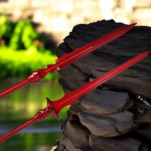 May include: Two red spears with sharp, pointed tips. The spears have intricate designs along the shaft and near the head. The background is blurred, with a natural setting of green foliage and dark wood.