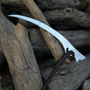May include: A silver scythe blade with a curved design and a dark brown handle. The handle has a textured, carved appearance. The scythe is set against a backdrop of weathered wooden logs, creating a rustic aesthetic.