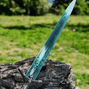 May include: A decorative sword with a teal blade featuring an intricate pattern. The hilt is silver with detailed carvings. The sword is displayed outdoors on a rock, with a blurred green background.