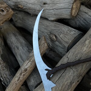May include: A curved, silver-colored scythe blade with a decorative handle. The blade has a sharp edge and a unique shape. The handle is dark brown with an ornate design. The scythe rests on a pile of weathered wooden logs.