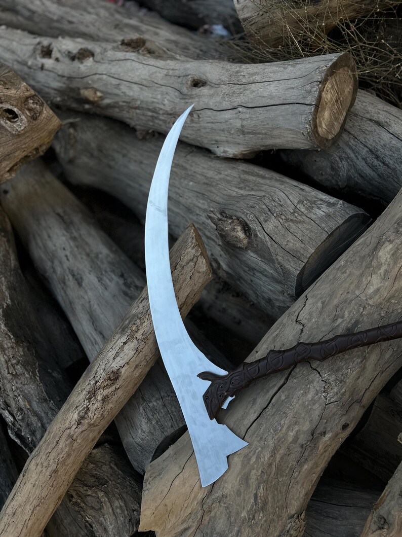 May include: A curved, silver-colored scythe blade with a decorative handle. The blade has a sharp edge and a unique shape. The handle is dark brown with an ornate design. The scythe rests on a pile of weathered wooden logs.