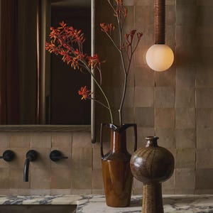 Peut inclure: Intérieur de salle de bain avec un vase en céramique marron et un vase rond plus petit sur une surface en marbre. Les vases contiennent des fleurs orange-rouge. Un luminaire mural avec une ampoule sphérique et un abat-jour marron texturé est également visible.