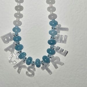 Op de afbeelding: Een kralenketting met doorschijnende witte en lichtblauwe ronde kralen. De ketting bevat witte letterkralen die het woord BACKSTREET vormen, waarbij de letters met kleine zilverkleurige metalen ringetjes in de kralenstreng zijn verwerkt.