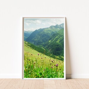以下が含まれることがあります： 鮮やかな緑の山々の風景を描いた縦型の額入りウォールアートプリント。前景には紫色の野花が点在する草地が広がり、背景には曇り空の下に丘陵や深い谷が広がっています。