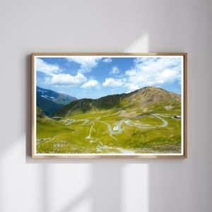 以下が含まれることがあります： 鮮やかな緑の草地を縫うように走る曲がりくねった山道と、白い雲が浮かぶ明るい青空が写る額装された風景写真。険しい地形、遠くの山頂、そして舗装された道がはっきりと見える、広大な山岳地帯の様子を捉えた一枚。