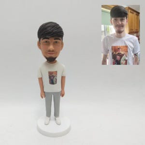 Könnte beinhalten: Eine Wackelkopf-Figur einer Person mit dunklem Haar und Bart, die ein weißes T-Shirt und eine graue Hose trägt. Das T-Shirt hat ein gedrucktes Bild. Die Figur steht auf einem weißen, kreisförmigen Sockel.