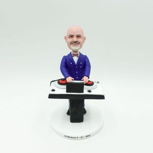 Puede incluir: Figura de un DJ con traje azul y pajarita, de pie detrás de una mesa de DJ blanca con tocadiscos rojos. La figura está sobre una base blanca con el texto "DJ Michael 9 - 27 - 2023".