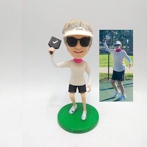Puede incluir: Figura de bobblehead de una persona con gafas de sol, visera y ropa deportiva, sosteniendo una paleta de pickleball. La figura está sobre una base verde con "G.O.A.T.". La imagen también incluye una foto de una persona jugando al pickleball.