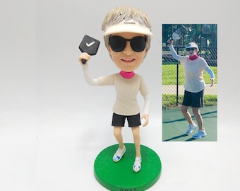 May include: A bobblehead figurine of a person wearing sunglasses, a visor, and athletic attire, holding a pickleball paddle. The figurine stands on a green base with the letters "G.O.A.T." The image also includes a photo of a person playing pickleball.