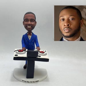 Puede incluir: Una figura de un DJ con traje azul, gafas y pajarita, detrás de una mesa de DJ con tocadiscos rojos y blancos. La base dice "PT by day, DJ by night". Una imagen de un hombre con traje está en el fondo.