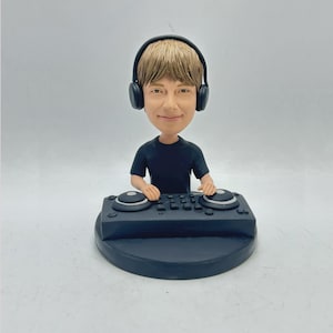Puede incluir: Figura de bobblehead de un DJ con auriculares y una camisa negra, de pie detrás de una mesa de mezclas negra. La figura tiene cabello castaño claro y está sonriendo. La base de la figura es redonda y negra.