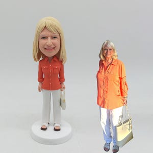 May include: A bobblehead figurine of a woman wearing an orange shirt and white pants, holding a white purse. Next to the figurine is a woman wearing a similar outfit, holding a white purse. Both are smiling.