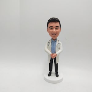 May include: A figurine of a smiling doctor wearing a white lab coat, blue shirt, black trousers, and black shoes. The doctor has a stethoscope and is standing on a white circular base.