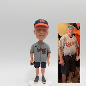 May include: A bobblehead figurine of a man wearing a gray baseball jersey with "Baltimore" and the number 35 in red. He wears a black and orange baseball cap, black shorts, and gray shoes. A smaller image of the man in a similar outfit is to the right.