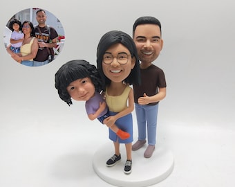 May include: A family figurine featuring a woman holding a child, with a man standing beside them. The woman wears a yellow top and blue skirt, the child a purple shirt and the man a brown shirt and blue pants. The figurine is on a white base.